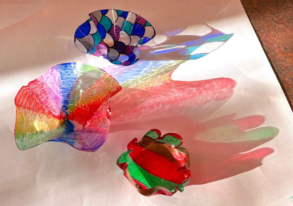 Melted plastic bowls shaped in the style of Dale Chihuly's glass bowls, by 8th grade students. Three bowls, one in vibrant shades of green and red, another painted with scalloped patterns of blue, turquoise, silver and pink, and the third with rainbow colors.